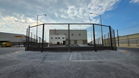 Outdoor sports court enclosed by tall black netting fence with street lights, surrounded by industrial buildings and paved ground under partly cloudy sky
