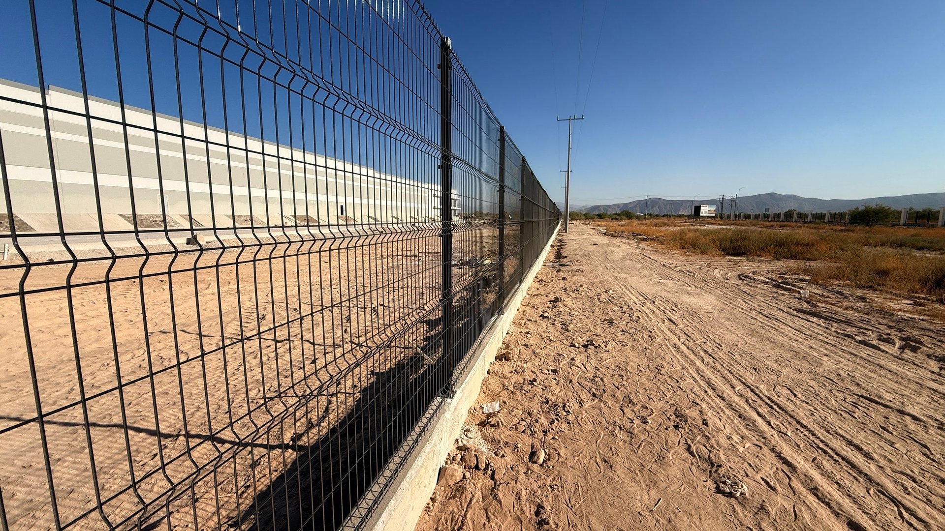 Metal mesh fence running along a desert dirt road under clear blue sky with mountains in the distance
