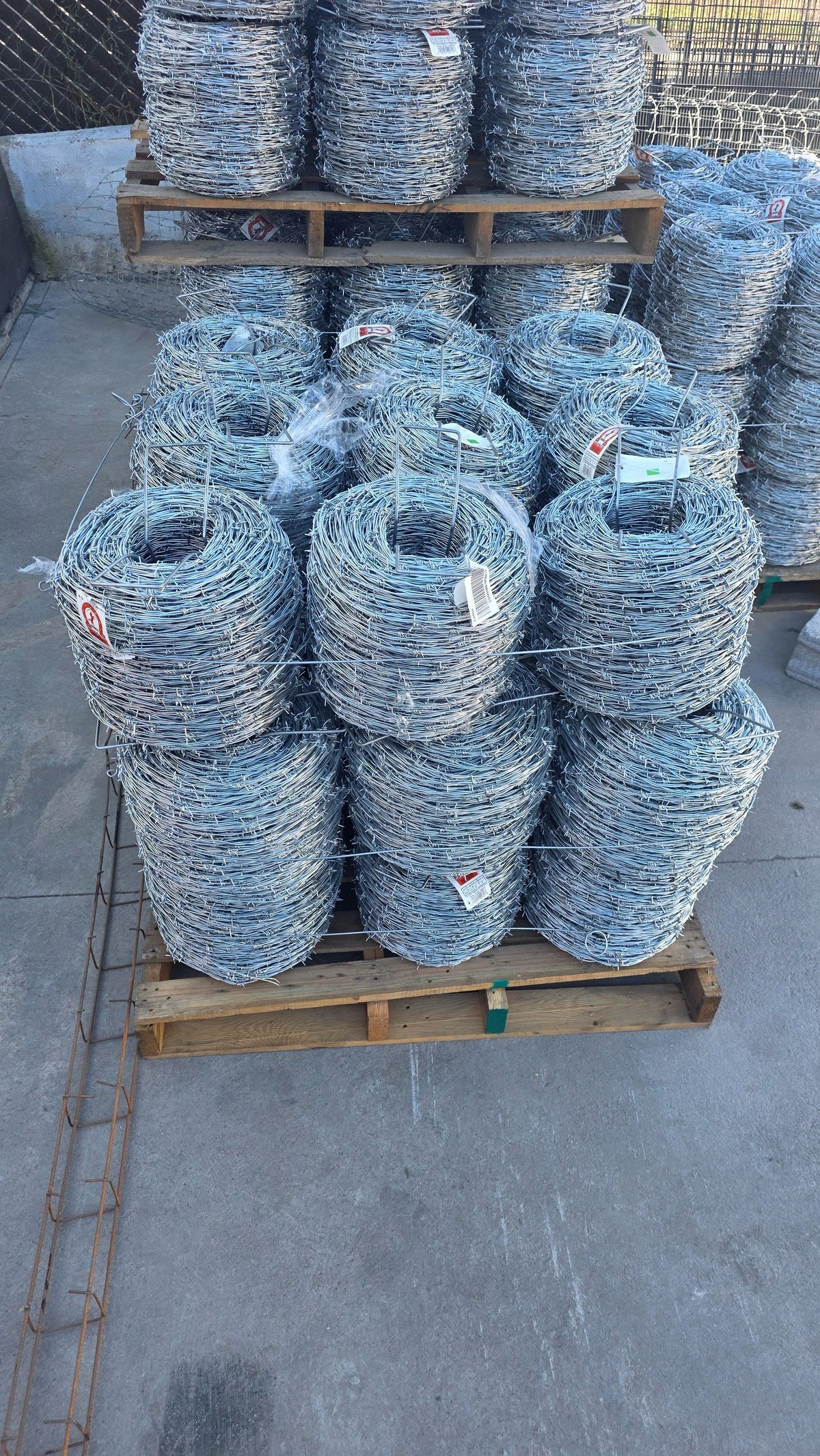 Stacked rolls of galvanized barbed wire on wooden pallets in an outdoor industrial storage area