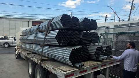 Truck loaded with stacked black metal pipes and tubes secured with straps in an industrial yard.