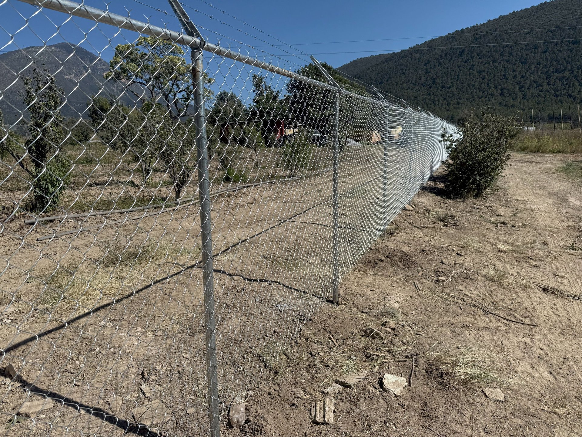 Chain-link fence enclosure in rural mountain valley with forested hills in background under clear blue sky