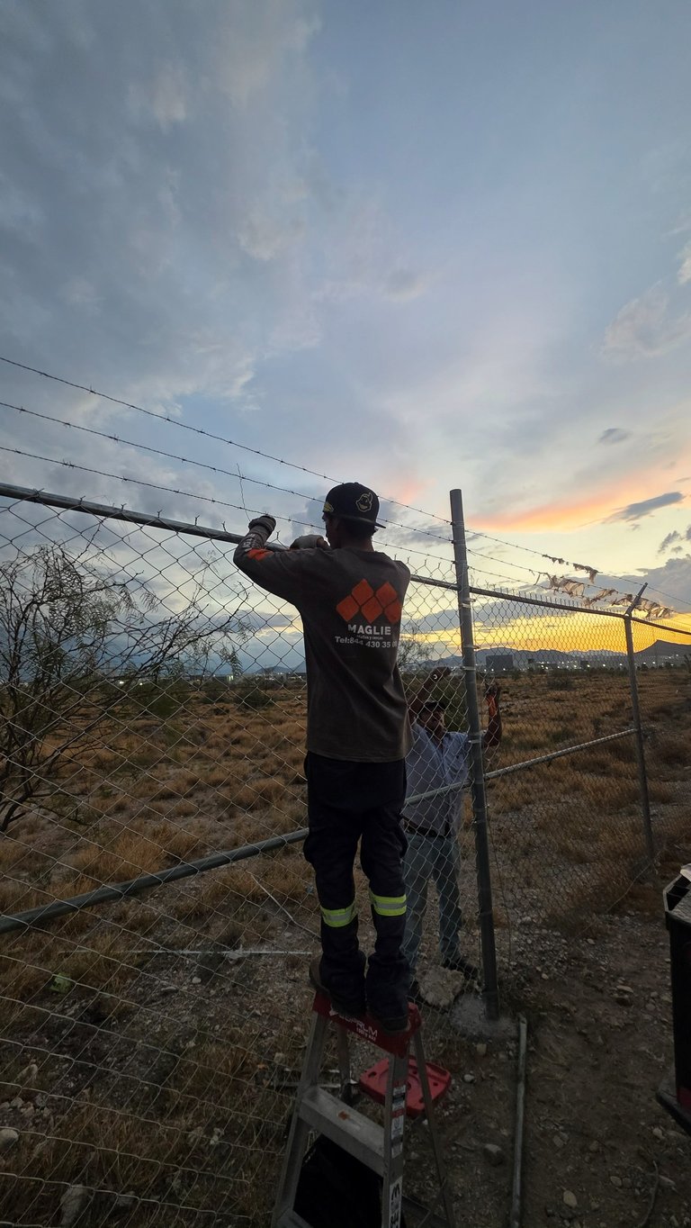 Person in dark jacket with reflective stripes standing on ladder at fence during sunset over rural landscape